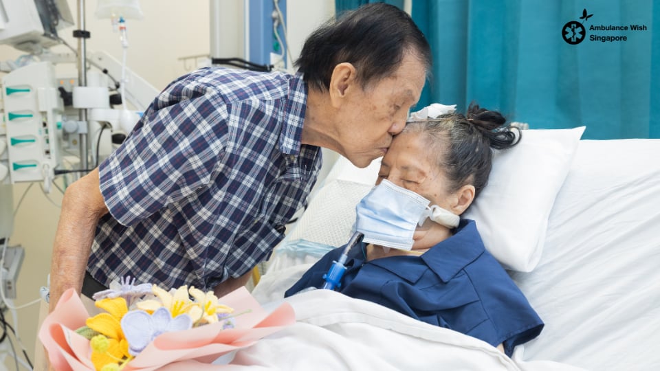 An elderly husband kisses his wife on the forehead at her hospital bedside, flowers beside them
