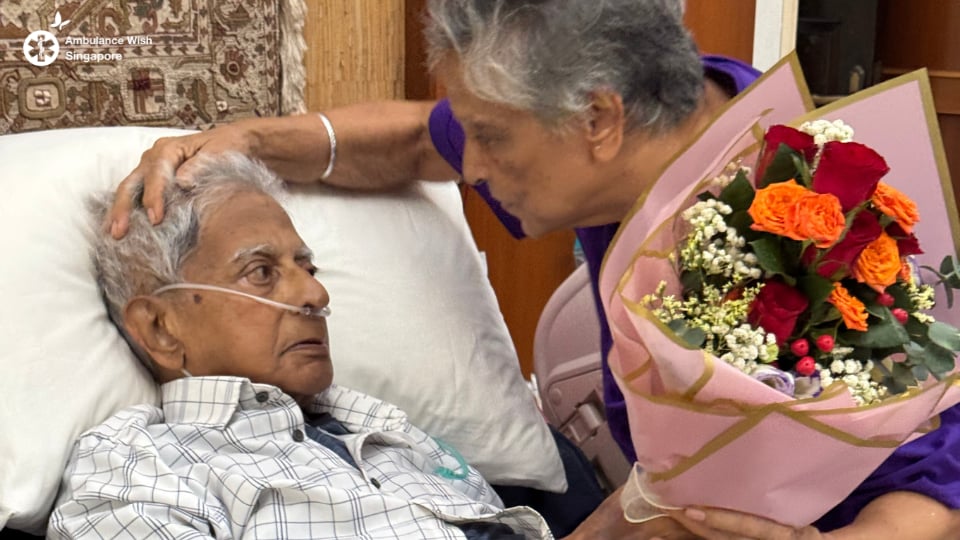 A wife leans close to her husband at his bedside at home, holding a bouquet of flowers
